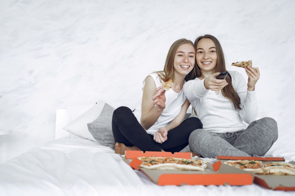 Two young women enjoying pizza and watching TV together indoors, sharing a joyful moment.