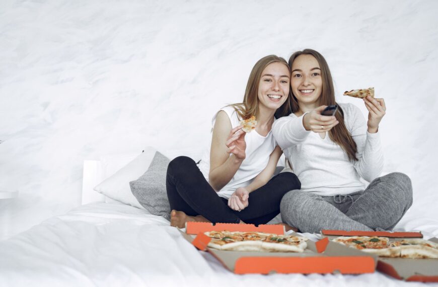 Two young women enjoying pizza and watching TV together indoors, sharing a joyful moment.
