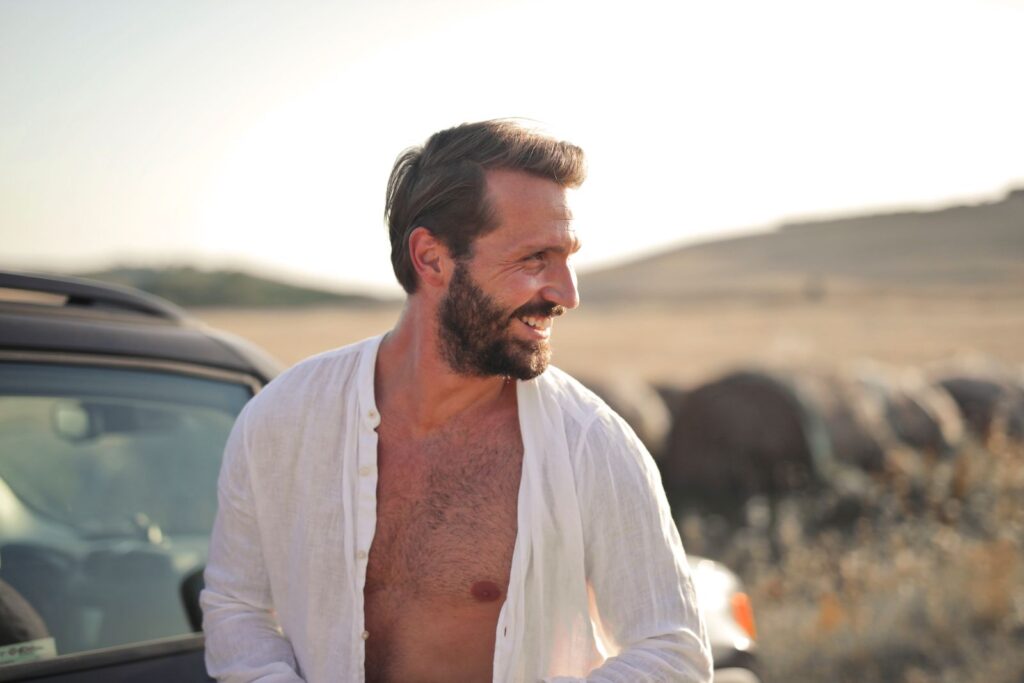 Bearded man smiling in open shirt, enjoying a sunny day outdoors next to a car.