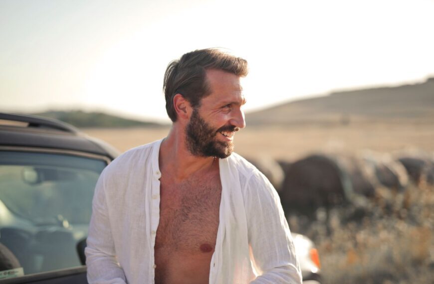 Bearded man smiling in open shirt, enjoying a sunny day outdoors next to a car.