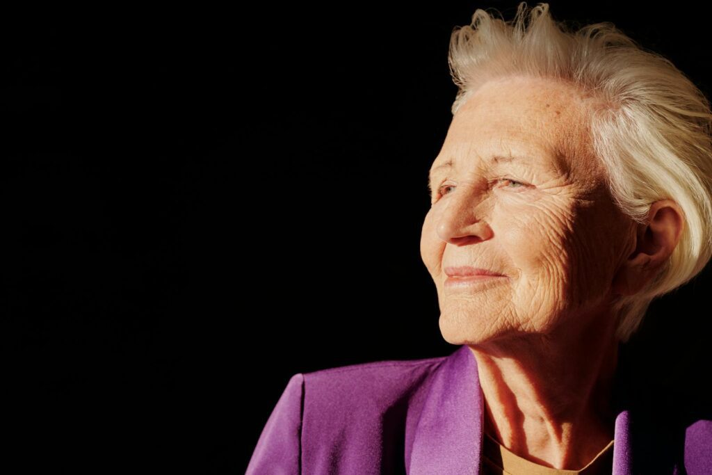 Elegant senior woman smiling with confidence in a purple jacket against black background.