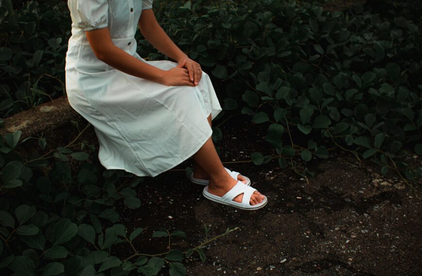 From above side view of crop unrecognizable female in trendy outfit and sandals sitting near shrubs in garden