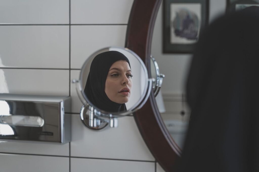 A woman wearing a hijab looks at her reflection in a bathroom mirror.