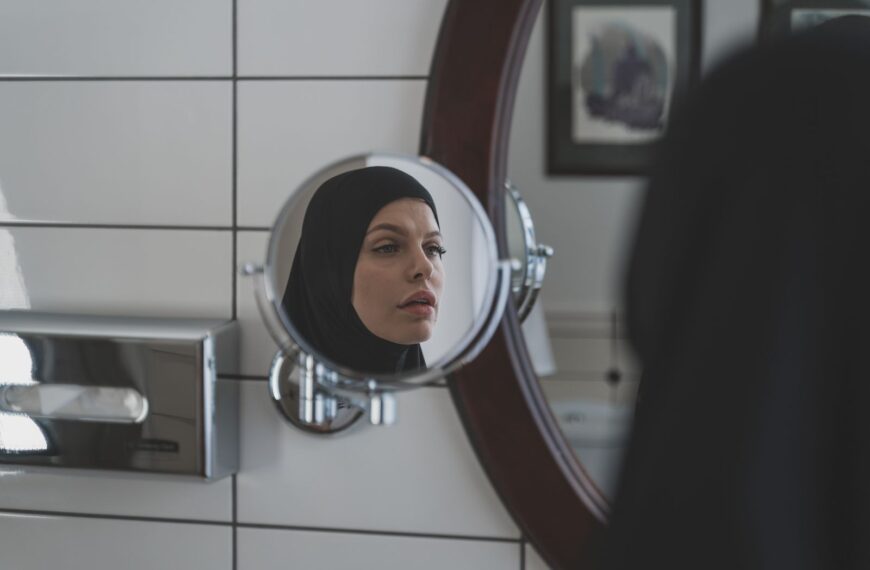 A woman wearing a hijab looks at her reflection in a bathroom mirror.
