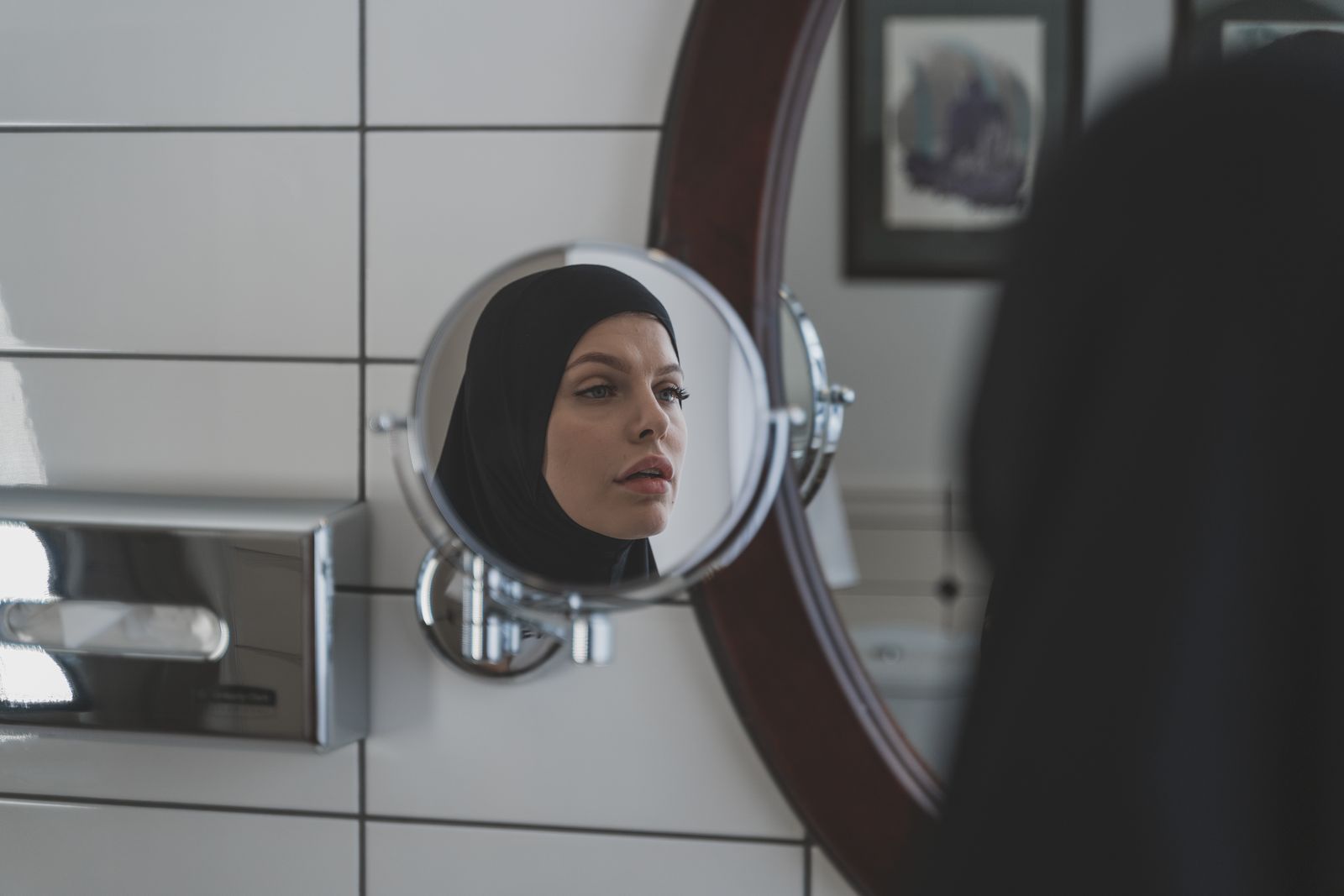A woman wearing a hijab looks at her reflection in a bathroom mirror.