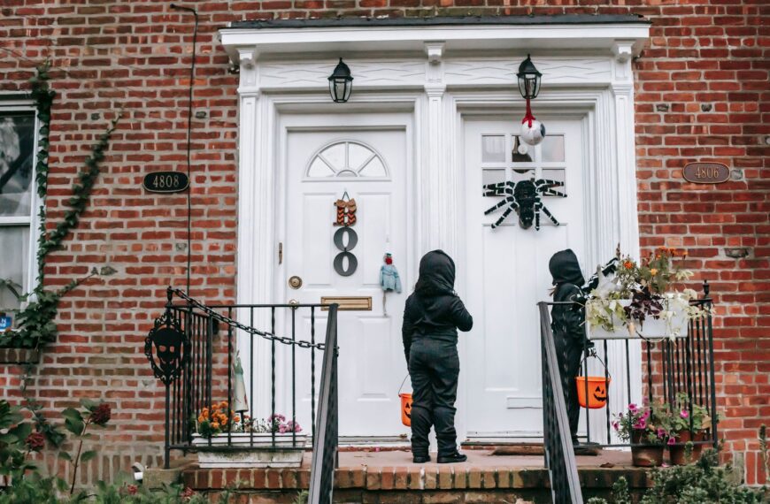 Full length of unrecognizable kids in Halloween costumes with masks standing near house door with buckets in daytime