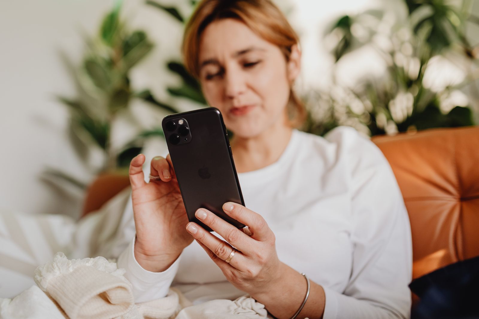 Woman comfortably sitting indoors using a smartphone with plants in the background.