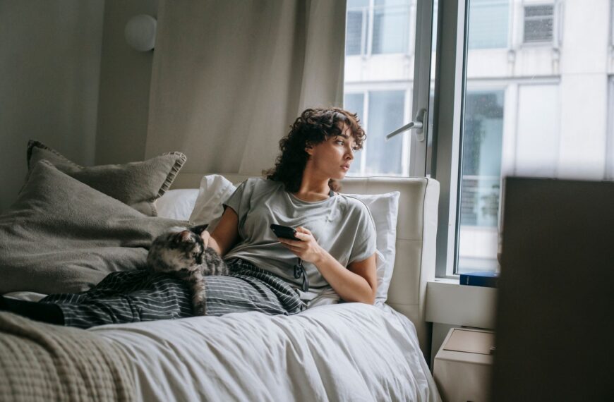 Full body of female owner looking at window and stroking cat while lying on bed and surfing smartphone on weekend