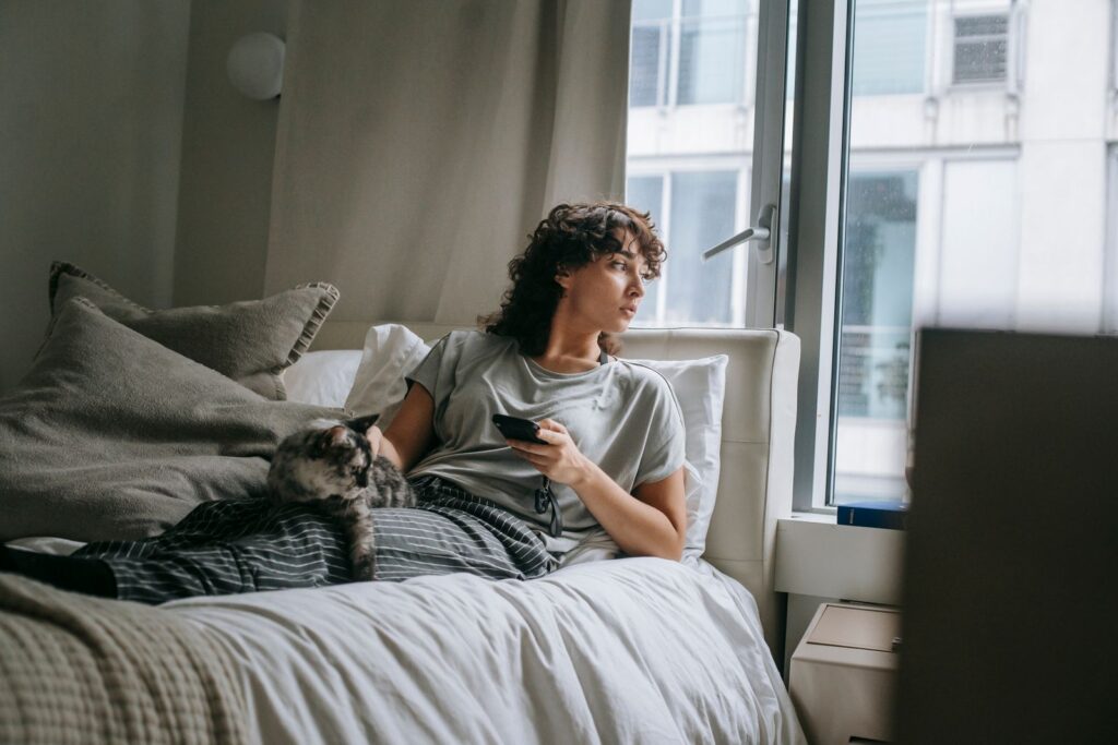 Full body of female owner looking at window and stroking cat while lying on bed and surfing smartphone on weekend
