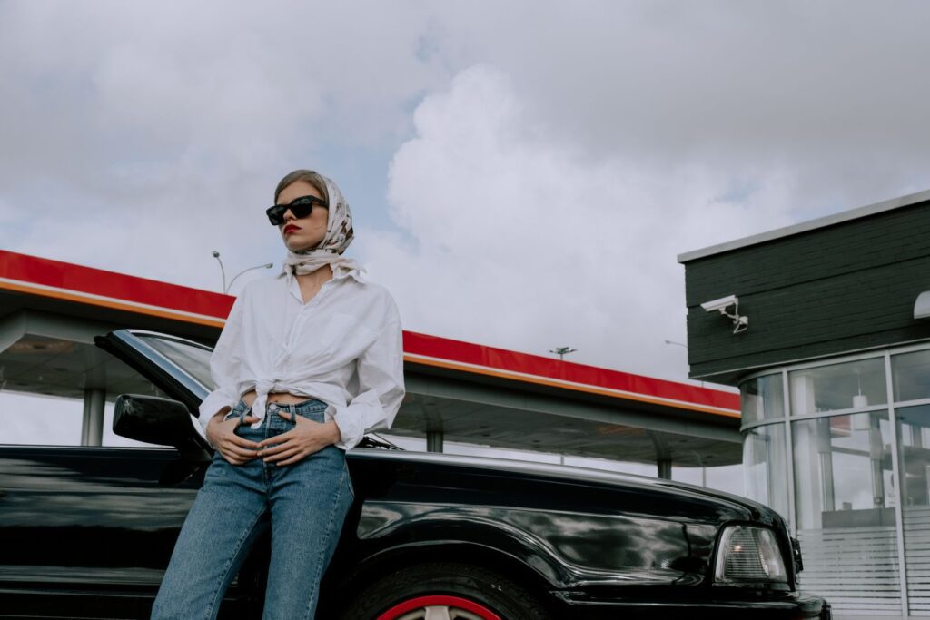 Fashionable woman in sunglasses leaning on a convertible at a gas station, exuding modern elegance.