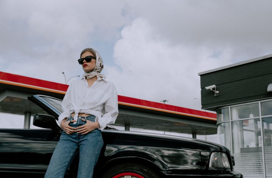 Fashionable woman in sunglasses leaning on a convertible at a gas station, exuding modern elegance.