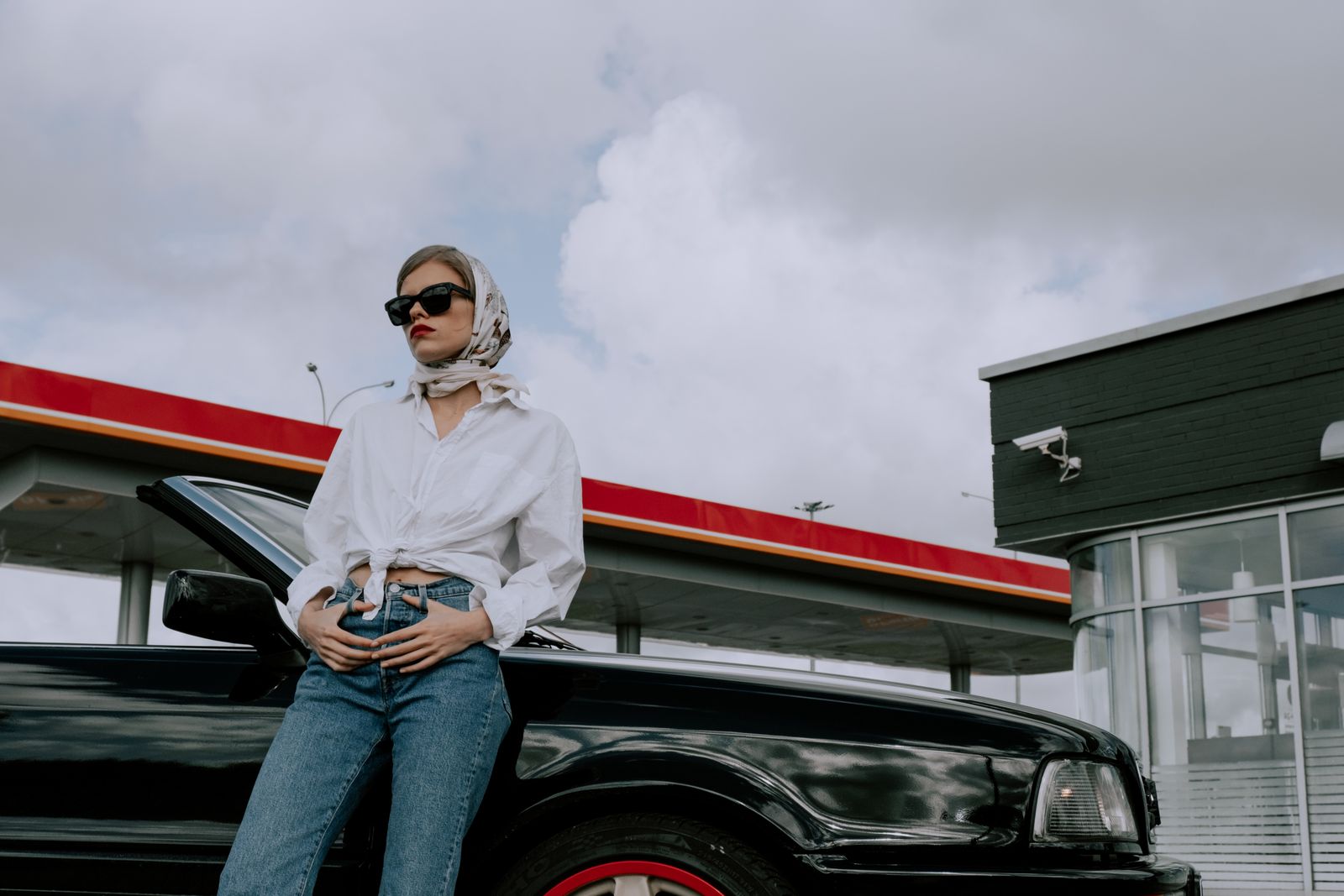 Fashionable woman in sunglasses leaning on a convertible at a gas station, exuding modern elegance.