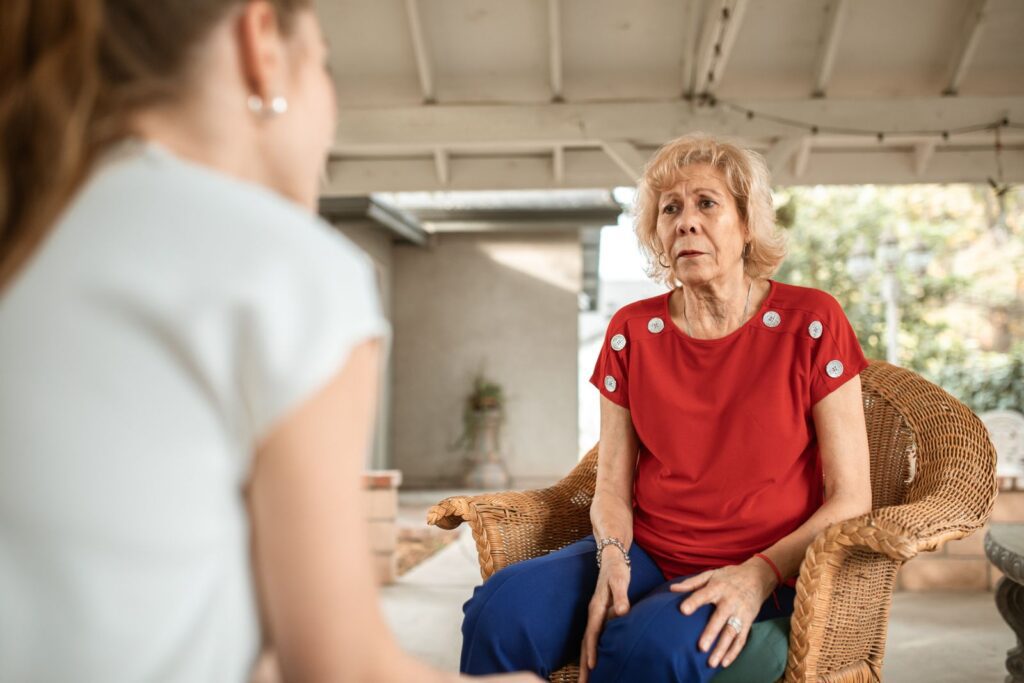 Senior woman in red shirt conversing indoors with younger woman. Daytime scene.