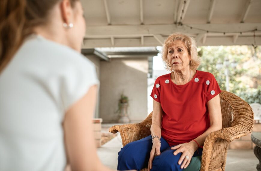 Senior woman in red shirt conversing indoors with younger woman. Daytime scene.
