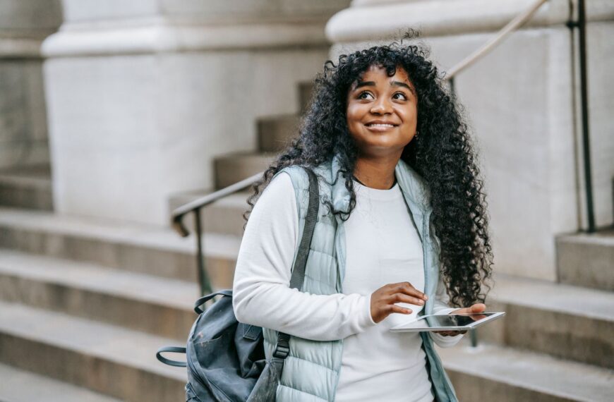 Happy young African American woman wearing casual clothes with backpack while using tablet on city street near building with stone stairs in daylight