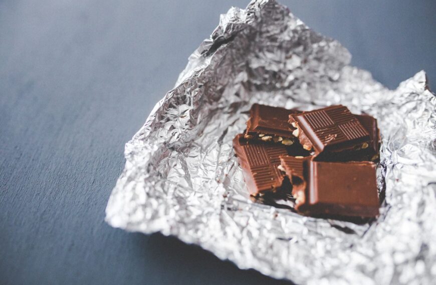 Close-up of a chocolate bar with nuts, partially unwrapped in foil, on a gray surface.