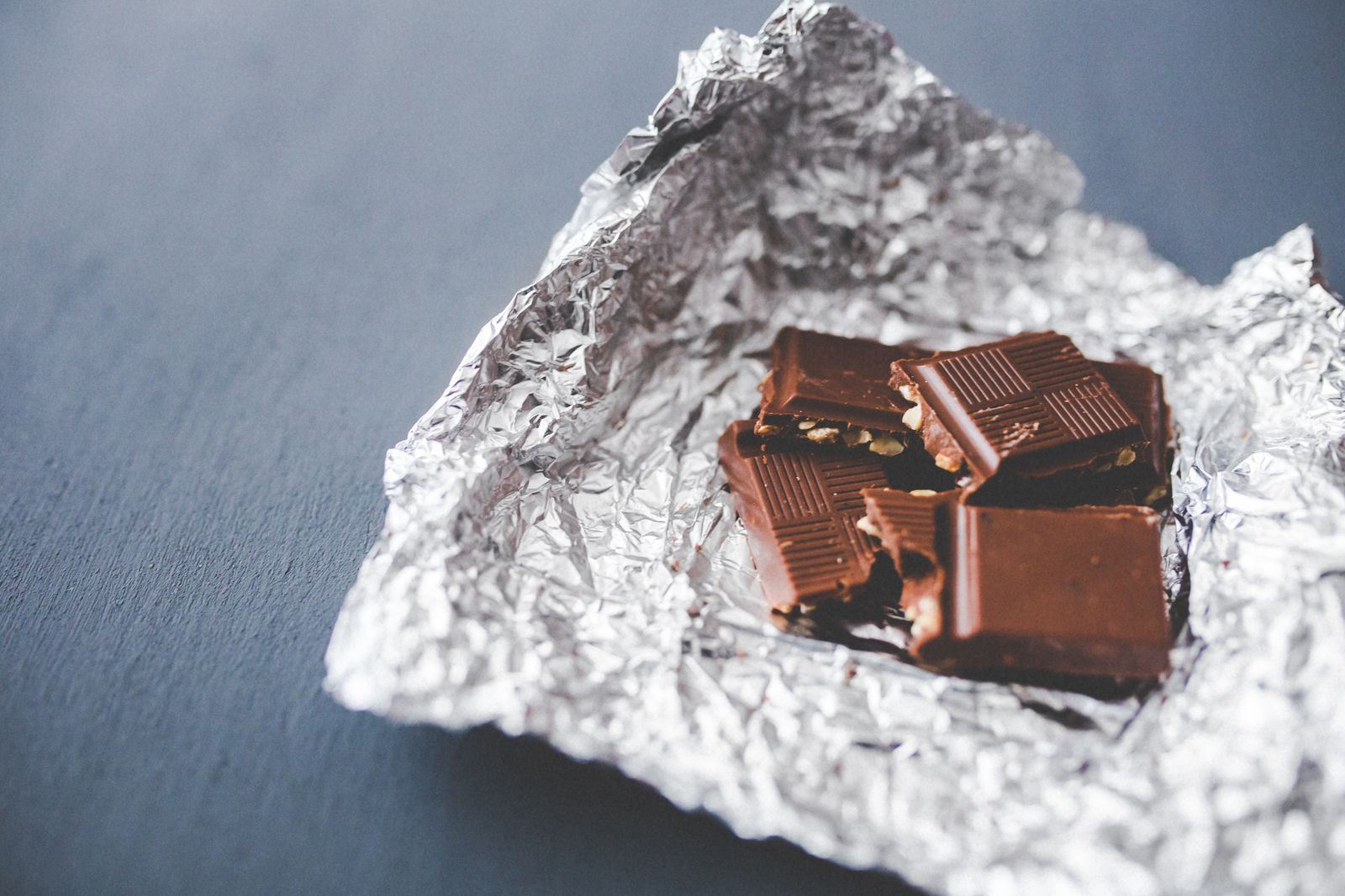 Close-up of a chocolate bar with nuts, partially unwrapped in foil, on a gray surface.