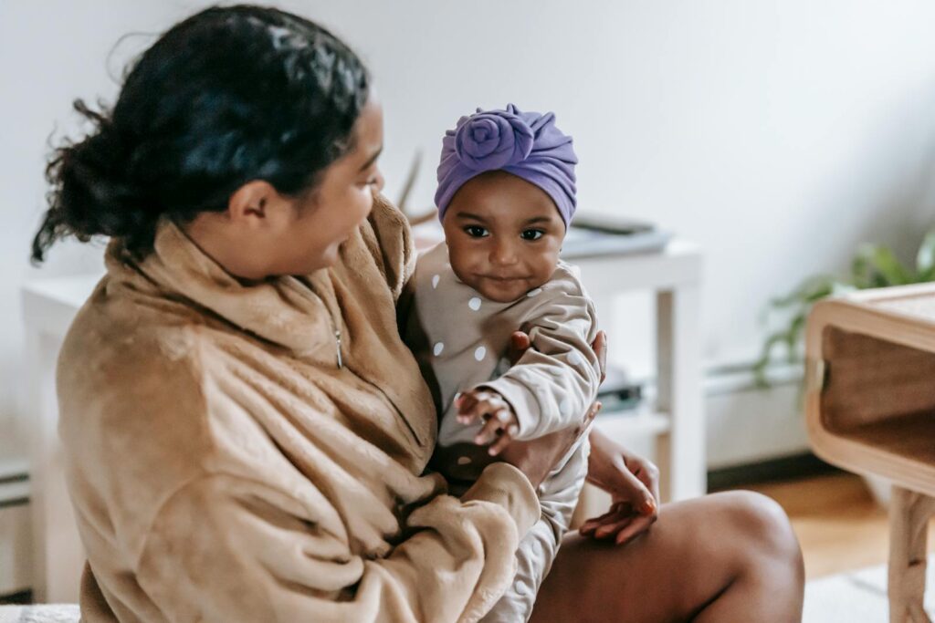 Positive African American mother looking at black little daughter on hands while spending time in living room on blurred background