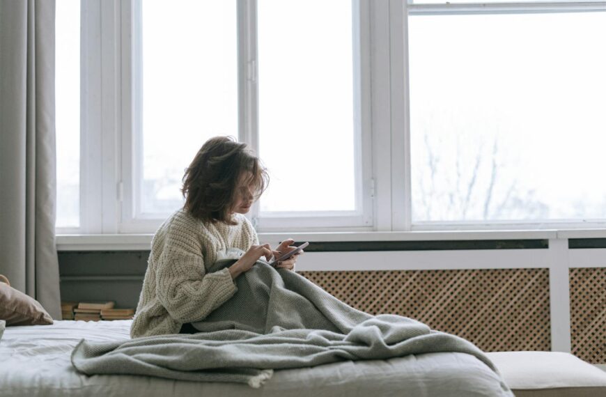 A woman in a knitted sweater using her smartphone while sitting on a bed with a blanket.