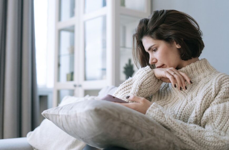 A woman in a knitted sweater using a smartphone, sitting pensively on a couch at home.