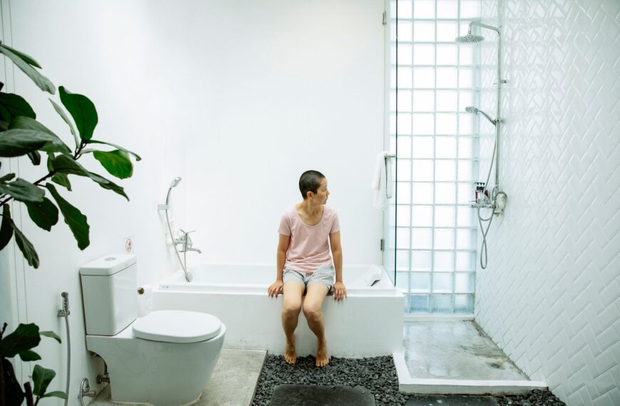 Full body of barefoot female in casual clothes sitting on bathtub next to toilet and shower in bathroom with green plant