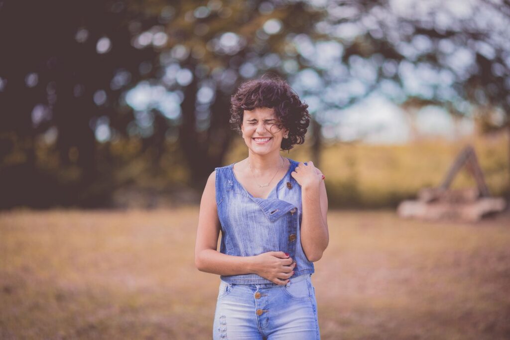 A happy woman with curly hair stands smiling in a sunny outdoor park setting.