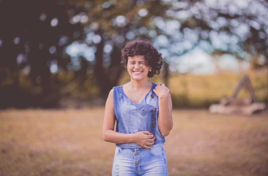 A happy woman with curly hair stands smiling in a sunny outdoor park setting.