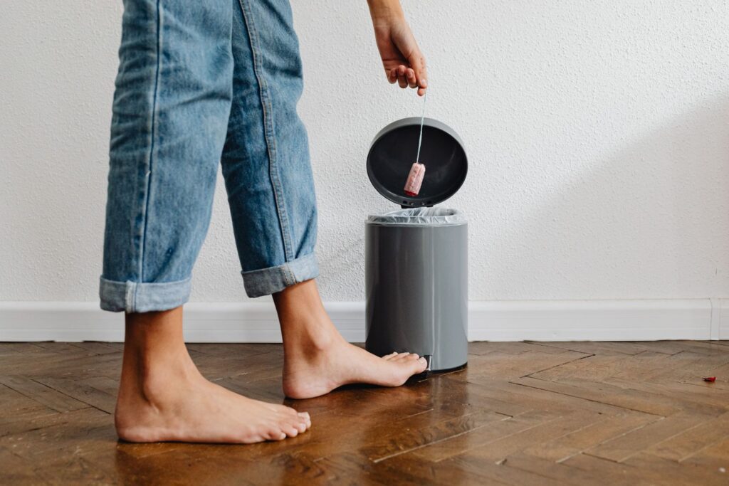 Barefoot woman in blue jeans discarding a tampon in a trash can at home, symbolizing menstrual care.