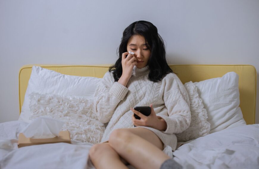 Asian woman in a sweater, looking sad and wiping tears, holding a smartphone seated on a bed.