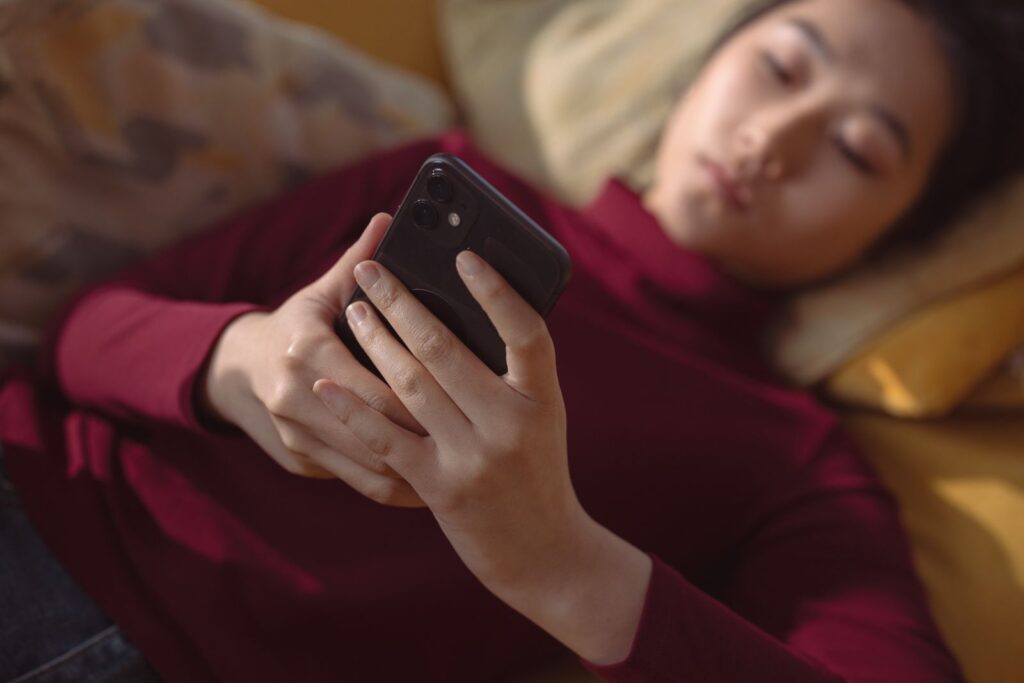 A young woman lying down, holding and using a smartphone for relaxation indoors.