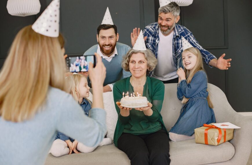 Grandmother holding cake, surrounded by family in party hats, smiling for a photo.