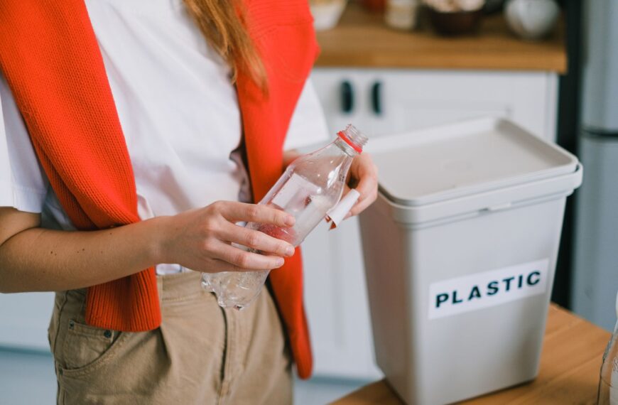 Woman placing plastic bottle into recycling bin in kitchen setting.