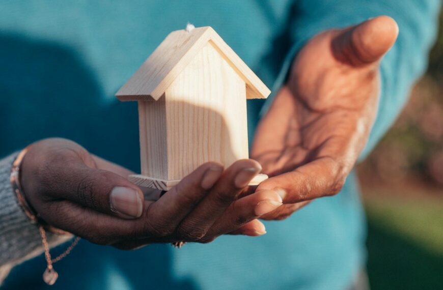 Close-up of hands holding a small wooden house model outdoors, symbolizing real estate investment.