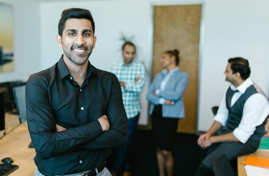A confident young professional standing in a modern office with colleagues interacting in the background.
