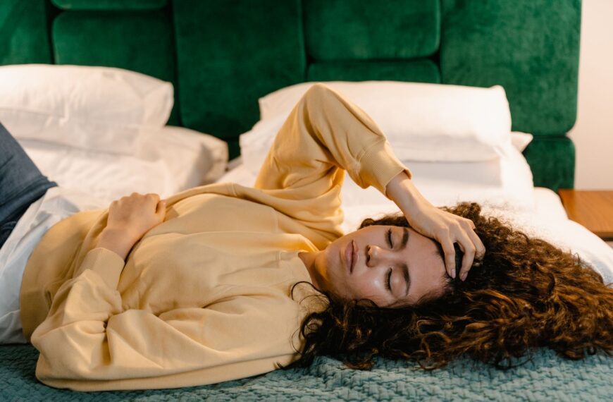 Woman lying on bed with hand on forehead, appearing relaxed or sleepy in a cozy indoor setting.