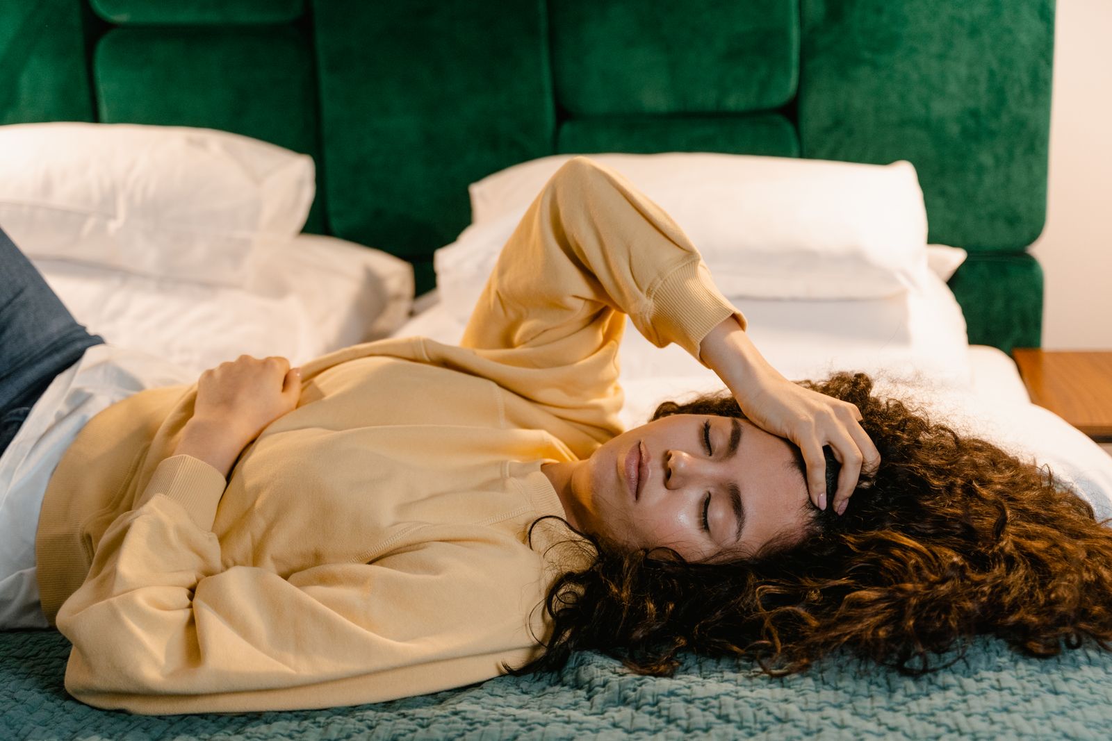 Woman lying on bed with hand on forehead, appearing relaxed or sleepy in a cozy indoor setting.