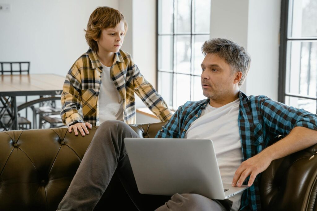 A father and son enjoying time together indoors, sharing a laptop on the sofa with a warm connection.