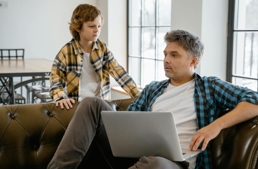 A father and son enjoying time together indoors, sharing a laptop on the sofa with a warm connection.