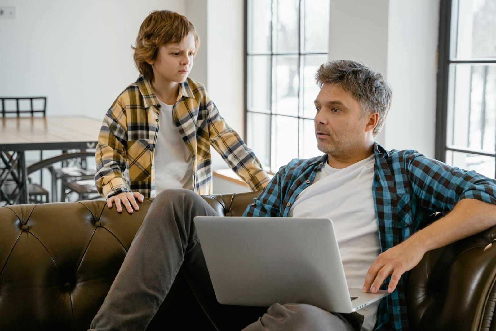 A father and son enjoying time together indoors, sharing a laptop on the sofa with a warm connection.