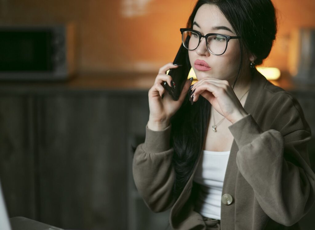 A woman with eyeglasses engaged in a phone call indoors, focused and thoughtful.