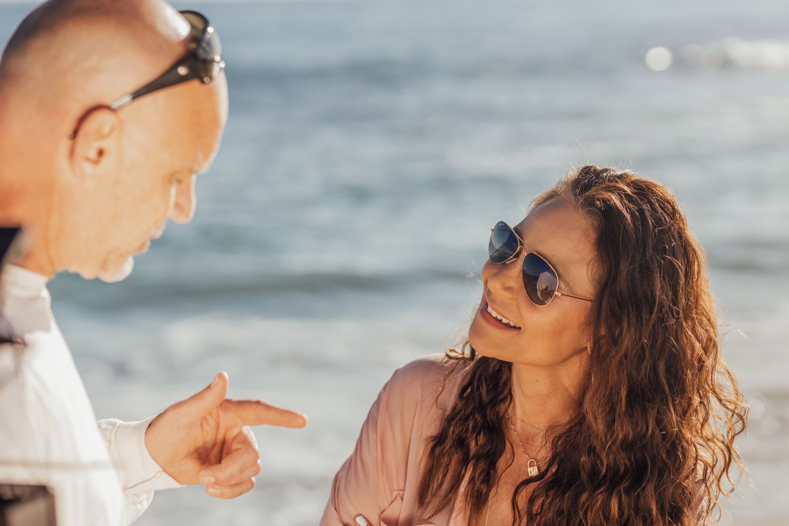A smiling couple enjoys a casual conversation by the ocean on a sunny day.