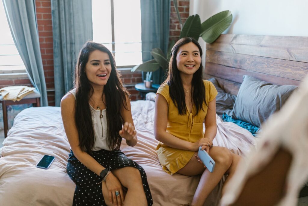 Two women enjoying a relaxing time together on a bed, sharing smiles and laughter in a bright, cozy bedroom.