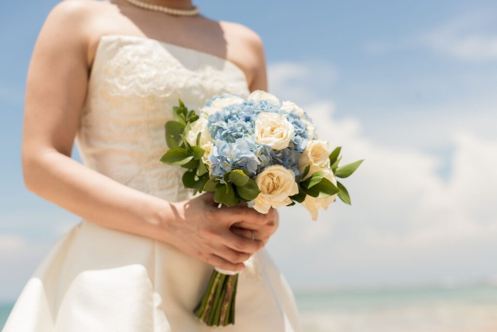 A bride holds a beautiful bouquet of blue hydrangeas and white roses on a sunny beach in Bali.