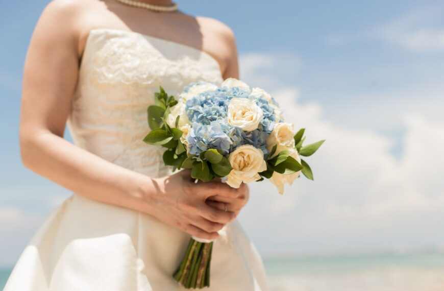 A bride holds a beautiful bouquet of blue hydrangeas and white roses on a sunny beach in Bali.
