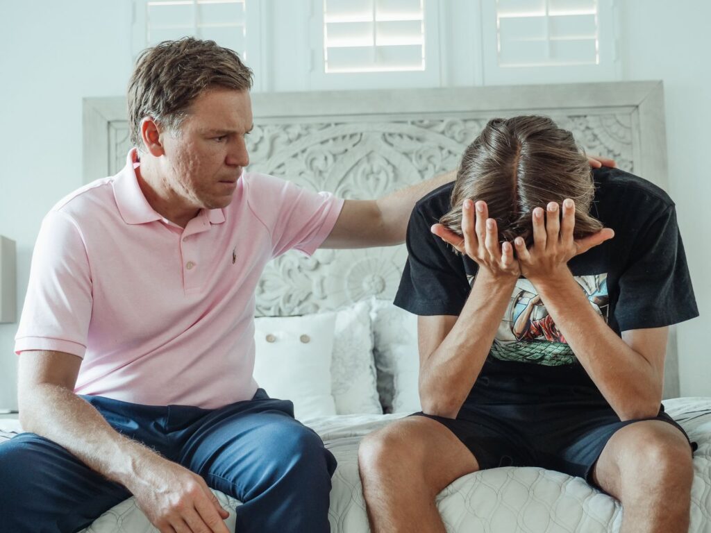 A compassionate father consoles his upset teenage son on a bed indoors.