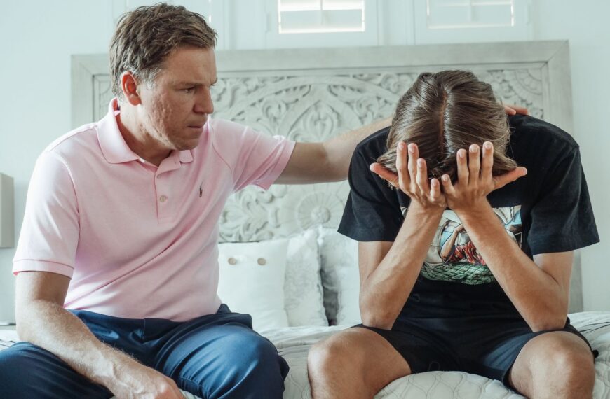 A compassionate father consoles his upset teenage son on a bed indoors.