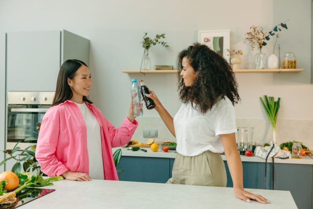 Two women in a kitchen clinking a soda bottle and water bottle, enjoying a healthy lifestyle moment.