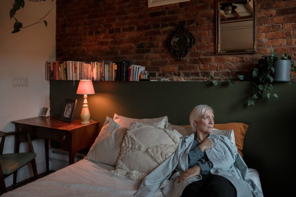 An elderly woman relaxes in a cozy, warmly lit bedroom with a brick wall backdrop.