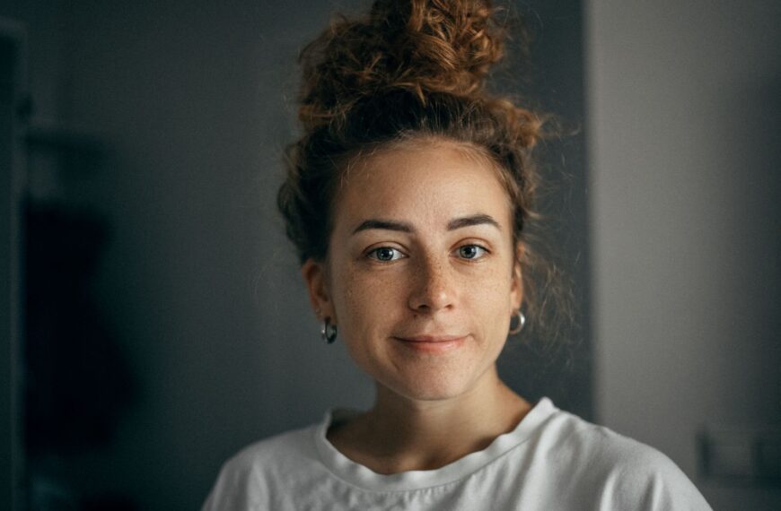 Portrait of a smiling woman with curly hair and freckles in soft light, wearing a white t-shirt indoors.