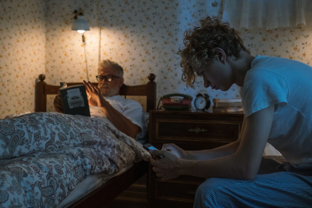 A serene scene of a grandfather reading a book while his grandson uses a smartphone in a cozy bedroom.